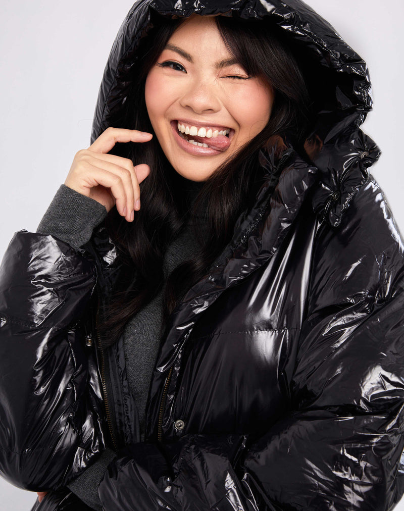 model has the hood up and is sticking her tongue out while wearing the lola black shine longline puffer jacket over a grey knit dress