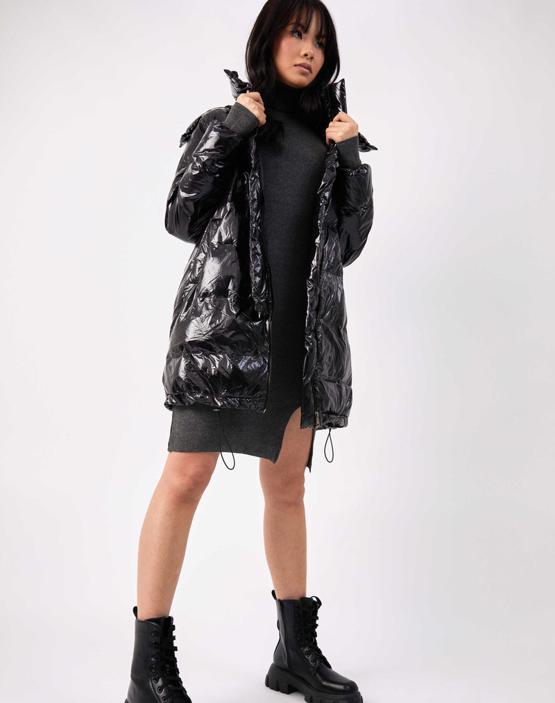 model is holding the lapels of the lola black shine longline puffer jacket over a grey knit dress and chunky boots