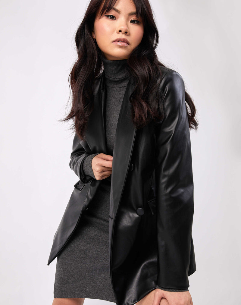 the model leans forward wearing the Rela Black Faux Leather Blazer over a knitted grey dress