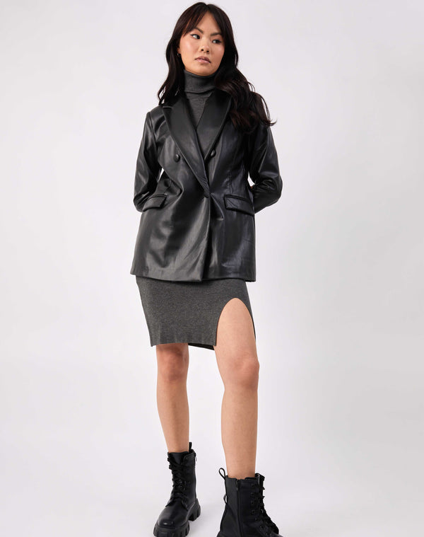 the model has her hands behind her back wearing the Rela Black Faux Leather Blazer buttoned over a grey knitted dress and black boots