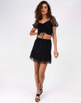 the model walks towards the camera while wearing the Olivia Black Ruched Front Mesh Crop and matching skirt with heels
