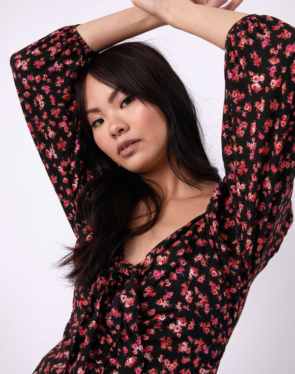 a close up shot of the models face with her arms above her head in the annie gathered tie front dress in a pink floral with black base in front of a white background