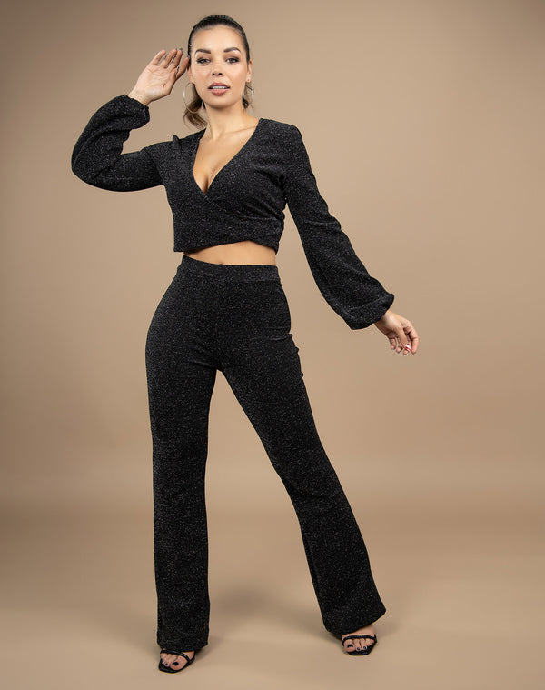 model wears black lurex two piece with wide trousers and wrap long sleeve top in front of a beige backdrop in a studio full length
