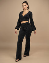 model wears black lurex two piece with wide trousers and wrap long sleeve top in front of a beige backdrop in a studio full