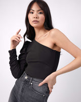 model has one hand raised to her face and her other on her hip the monet black one shoulder bodysuit with grey jeans
