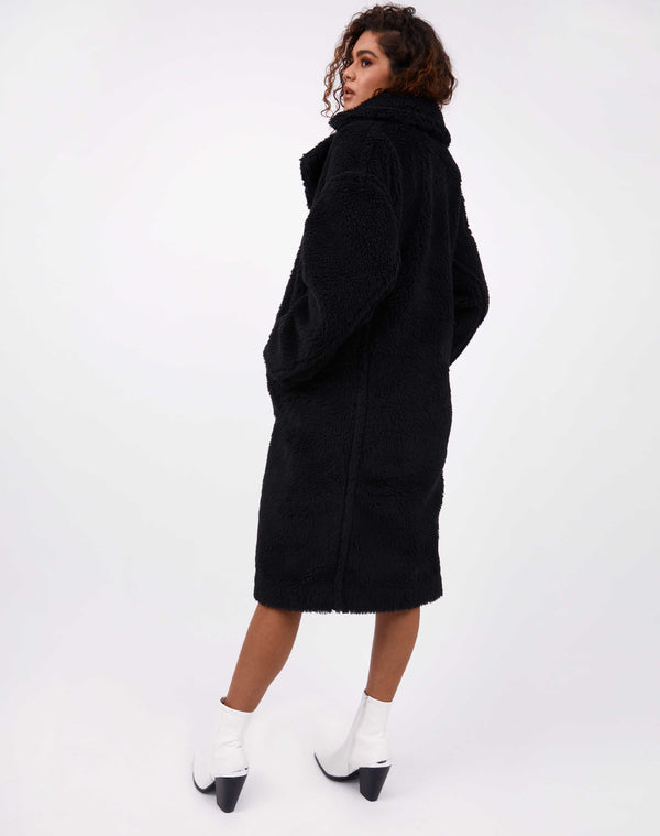 model faces away from the camera in gigi black teddy longline coat with white boots