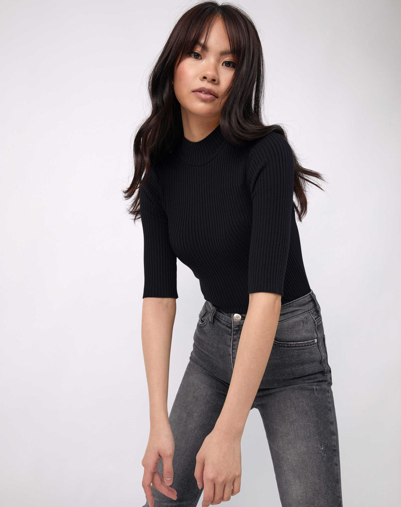 model leans towards the camera the meg ribbed mid sleeve bodysuit in black with grey jeans
