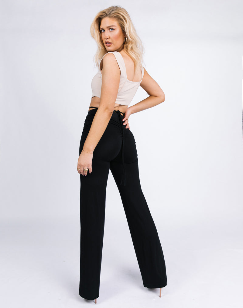 model turns to show the back of the florence ruched back trousers in black with tie details with nude heels and a beige corset