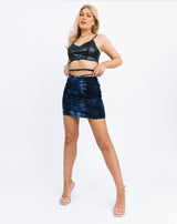 the model holds the tie details of the Remi Blue Tie Dye Ruched Skirt in a full length image wearing a pu crop top and heels